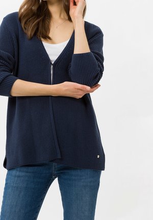 Kobieta ubrana w granatowy dzianinowy kardigan na białej bluzce i niebieskich dżinsach, z jedną ręką skrzyżowaną na piersi, a drugą dotykającą twarzy.