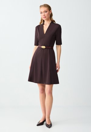 Femme debout portant une robe marron foncé longueur genoux avec manches mi-longues, décolleté en V et ceinture marron avec boucle dorée, accompagnée de talons foncés.