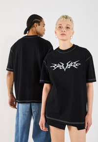 Black oversized t-shirts with white graphic "SAVAGE" design, contrast stitching, short sleeves, and relaxed fit, modeled by two individuals.