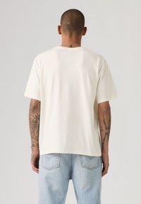 T-shirt en coton blanc, coupe décontractée, manches courtes et col rond, présenté de dos. Montre des tatouages visibles sur les bras.