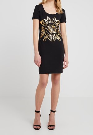 Robe noire à manches courtes avec un tigre ornemental doré et le logo « Versace Jeans », portée avec des sandales à talons hauts noires à bride de cheville sur un fond uni.