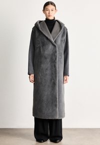 Sivý dlhý kabát vyrobený z mäkkého, textúrovaného materiálu. Má kapucňu, otvorený predok a voľný strih, štýlovo skombinovaný s čiernymi nohavicami s širokými nohavicami.