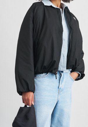 Personne portant une veste bomber noire courte par-dessus une chemise boutonnée bleu clair et un jean bleu clair, tenant une casquette noire dans une main.