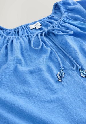 Modrá blůza vyrobená z texturovan é tkaniny, se strečovým výstřihem a šňůrkou se třásněmi z korálků. Štítek ukazuje značku "NEXT."