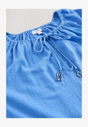 Modrá blůza vyrobená z texturované tkaniny, se strečovým výstřihem a šňůrkou se třásněmi z korálků. Štítek ukazuje značku "NEXT."