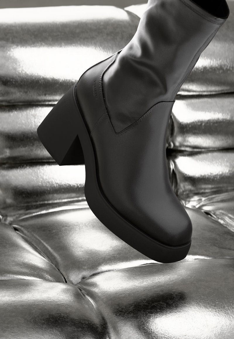 Bottine noire en cuir avec un talon épais, présentant une forme élégante et une texture lisse, posée sur une surface métallique brillante.