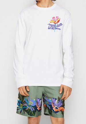 Person wearing white long-sleeve shirt with "Amsterdam Wildflowers" and flower graphic, paired with green shorts featuring large colorful floral prints.