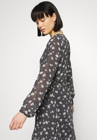 Vestido estampado floral em tecido preto transparente com flores brancas e amarelas, mangas bufantes longas e cintura ajustada, apresentando um ajuste solto.