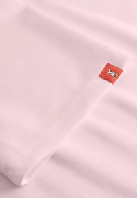 Jasnoróżowa tkanina z podwiniętym brzegiem, na którym znajduje się mała czerwona metka haftowana białym symbolem dłoni blisko szwu.