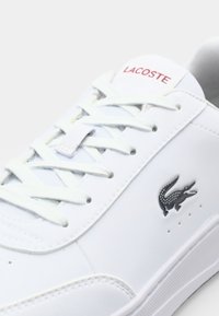 Baskets blanches Lacoste avec lacets blancs, logo Lacoste rouge sur la languette et emblème crocodile noir sur le côté.