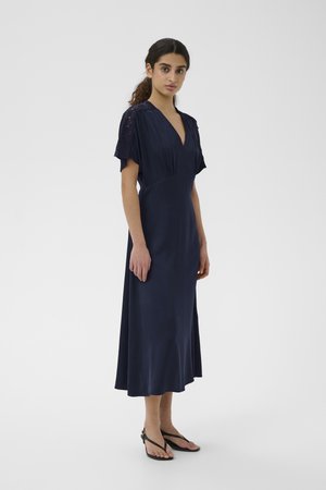 Femme aux cheveux foncés en queue de cheval basse portant une robe midi bleu marine avec des manches courtes en dentelle et des sandales noires à brides, debout sur un fond uni.