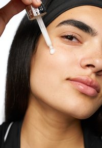 Un siero trasparente viene erogato da un contagocce sulla guancia di una persona con tono della pelle medio, pelle liscia e capelli scuri e lisci.