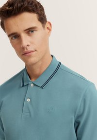 Polo shirt azzurro chiaro con due bottoni, colletto a righe blu navy e logo ricamato sottile sul petto. Tessuto dalla superficie liscia.