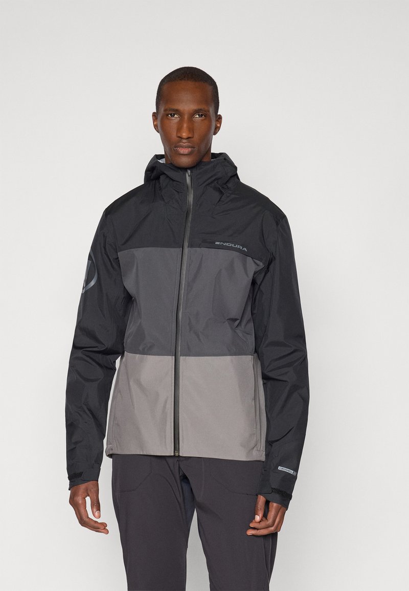 Veste étanche en coloris noir et gris avec capuche, fermeture éclair et logo sur la poitrine, présentant une coupe structurée et des poignets élastiques.