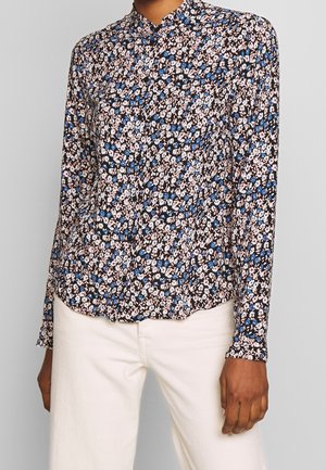 Kvinde iført en sort bluse med lange ærmer med små pink og blå blomsterprint, kombineret med hvide bukser med høj talje.