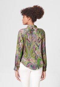 Langarmshirt in een levendige groene, paarse en gouden paisleypatroon. Zachte stof met een kraag en knoopmanchetten, gestyled met een witte broek.