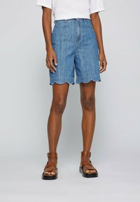 Ljust blå denimshorts med scalloped fåll, hög midja, femfickorsdesign och slät front, tillsammans med bruna spännremssandaler.