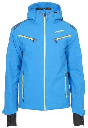 Mann in blauem und rotem Skianzug mit Reißverschlusstaschen, steht mit einer Hand in der Tasche vor einfarbigem Hintergrund.