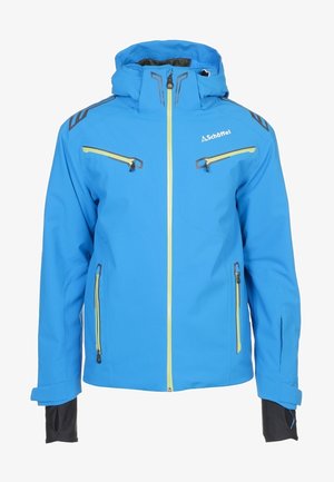 Homme portant une combinaison de ski bleue et rouge avec des poches zippées, debout avec une main dans la poche devant un fond uni.