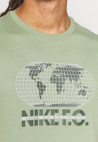 Camiseta verde claro con un gráfico de un globo blanco con líneas de cuadrícula y continentes verde oscuro. Texto "NIKE F.O." mostrado de manera destacada debajo.