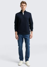 Sudadera con cremallera azul marino con un patrón texturizado, puños y dobladillo de canalé, combinada con jeans azules y zapatillas blancas, de pie contra un fondo claro.