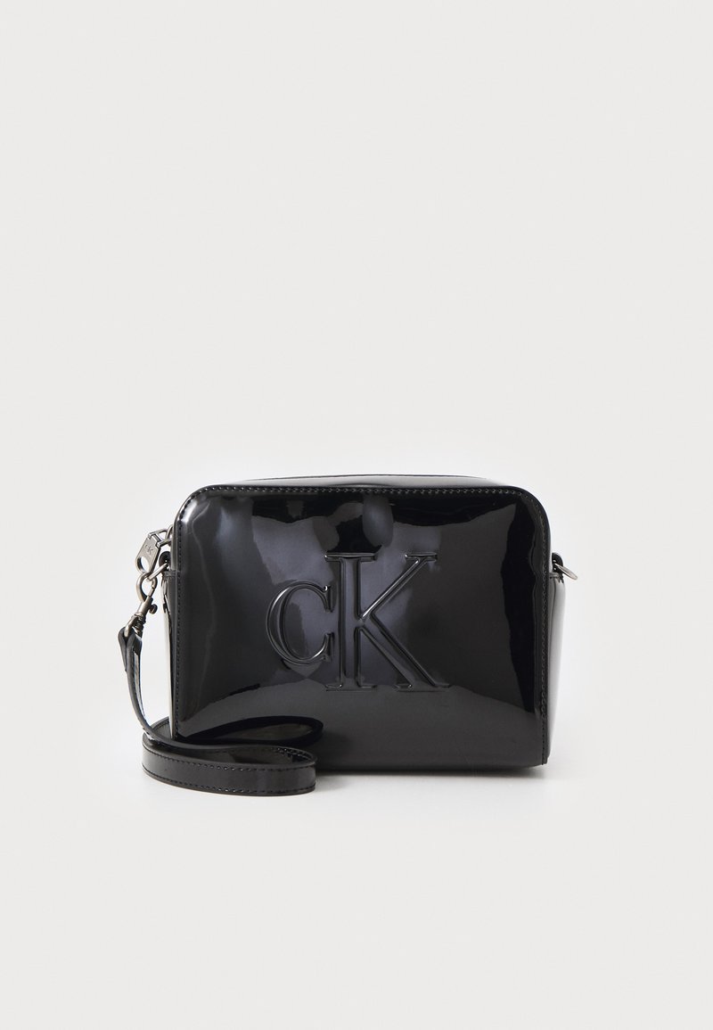 Svart lakkert lær crossbody-veske med preget "CK"-logo, rektangulær form, glidelås på toppen og avtakbar stropp. Elegant, blank finish.