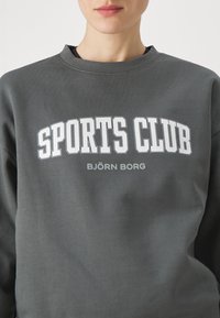 Felpa grigia in cotone con collo rotondo. Presenta una scritta grande in bianco "SPORTS CLUB" e "BJÖRN BORG" stampata sulla parte anteriore.