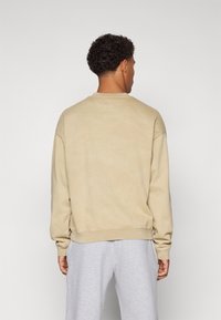 Beige színű, lágy anyagból készült crewneck pulóver, laza fitnesz kialakítással, bordás mandzsettákkal és aljjal. Világosszürke melegítőnadrággal viselve.