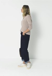 Světle béžový svetr s dlouhými rukávy, spárovaný s tmavě modrými džíny s manžetami. Bílé tenisky s béžovými akcenty. Neutrální pozadí.