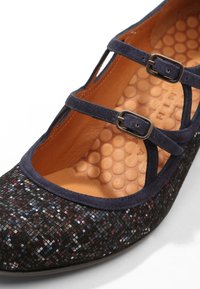 Chaussure noire à motif multicolore avec bout rond, dotée de deux sangles bleu marine, boucles bronze et une semelle intérieure orange texturée.