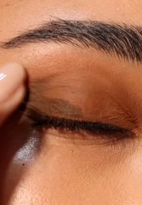 Close-up van een gesloten oog met een lichtbruine ooglid, contrasterende donkere wimpers en een vingertip die voorzichtig het ooglid omhoog trekt.