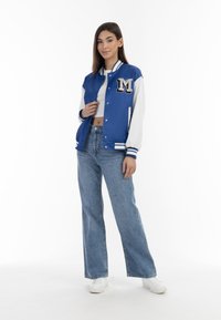 Giacca varsity blu e bianca con un patch a forma di "M", abbinata a jeans a gamba larga blu chiaro e sneakers bianche, indossata da un modello su sfondo bianco.