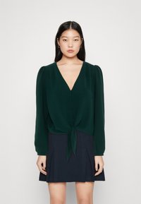 Dark green blouse with a V-neck, long puff sleeves, and button details, tied at the front over a navy pleated skirt. Smooth texture.
