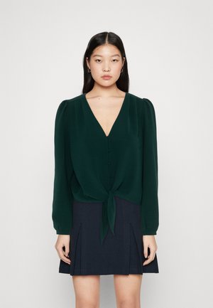 Dark green blouse with a V-neck, long puff sleeves, and button details, tied at the front over a navy pleated skirt. Smooth texture.