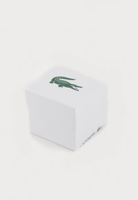 Weißer Karton mit strukturierter Oberfläche, oben mit einem grünen Krokodil-Logo und seitlich mit dem Text "LACOSTE".
