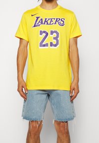 Man som bär en gul Lakers t-shirt med nummer 23 i lila och ljusblå slitna jeansshorts, stående mot en enfärgad bakgrund.