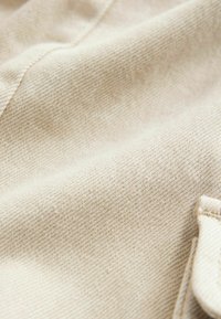 Tissu en denim beige avec une texture lisse, présentant un motif de tissage subtil. Une petite poche est partiellement visible sur le bord.