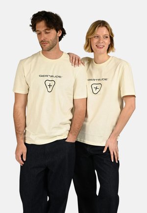 Un homme et une femme portant des t-shirts beige assortis « GERTRUDE » et des pantalons foncés, la femme posant sa main sur l'épaule de l'homme, souriante.