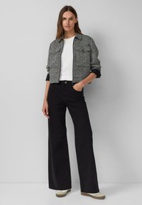 Kobieta w czarnych szerokich spodniach, białych trampkach, białym t-shircie i skróconej, wzorzystej czarno-białej kurtce stoi przy gładkiej ścianie.