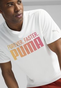 Balta trumpa rankovėmis sportinė marškinėliai su tekstūruotu audiniu, su daugiaspalviu gradientiniu tekstu: "AMŽINAI. GREIČIAU. PUMA," ir logotipo akcentu.