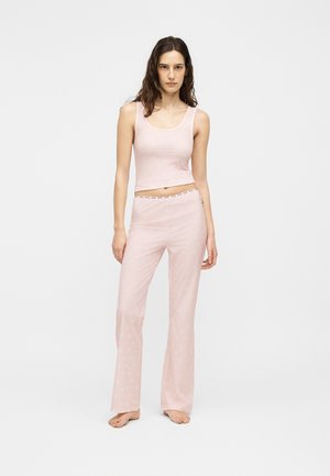 Jeune femme aux longs cheveux foncés, debout pieds nus, portant un crop top sans manches rose clair et un pantalon ample assorti avec des motifs subtils.