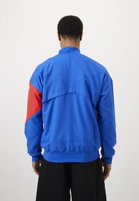 Blue jacket with red accents, featuring a high collar, elastic cuffs, and a distinctive back design. Made of smooth, lightweight fabric.