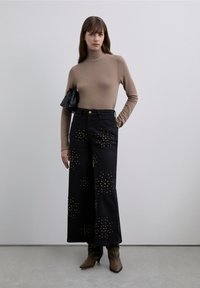 Vaqueros negros de pierna ancha con adornos dorados con tachuelas, combinados con un suéter ajustado de cuello alto marrón y un pequeño bolso negro.