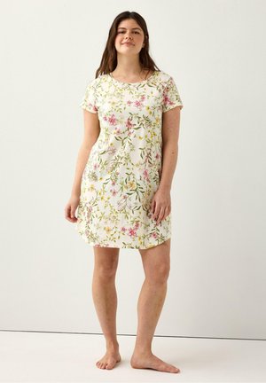 Donna in piedi a piedi nudi che indossa un vestito bianco a maniche corte, lungo fino al ginocchio, con motivi floreali rosa, gialli e verdi su uno sfondo neutro.