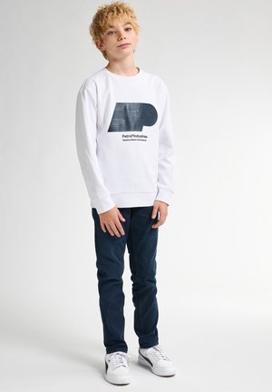 Witte sweatshirt met een blauw grafisch logo en tekst, gecombineerd met donkerblauwe jeans en witte sneakers. Model staat tegen een neutrale achtergrond.