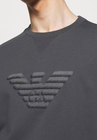 Mörkgrå sweatshirt med en upphöjd, tonad logotyp och horisontella ränder. Halsringningen är i crew-stil med ribbad struktur.