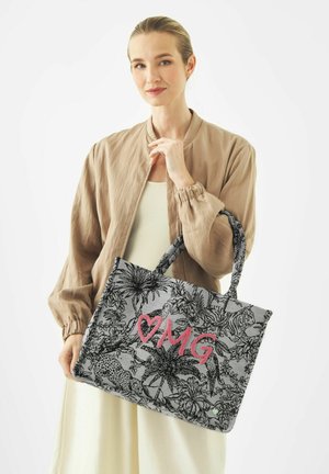 Vrouw met blond haar in een beige jasje die een grijze tas vasthoudt met zwarte jungle-dieren en roze "OMG"-tekst.