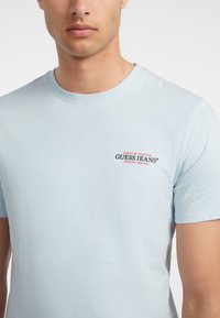 Tricou din bumbac de culoare bleu deschis cu guler rotund, având textul logo "GUESS JEANS" în roșu și negru pe partea stângă a pieptului.