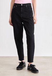 Melni džinsi ar augstu jostasvietu un sašaurinātu piegriezumu, ar divām priekšējām kabatām un pogas aizdari, kombinēti ar melniem sporta apaviem.