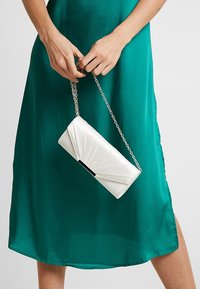 Pochette blanche au design plissé, avec une chaîne argentée et une touche noire. Portée avec une robe en satin verte, mettant en valeur sa silhouette élégante.